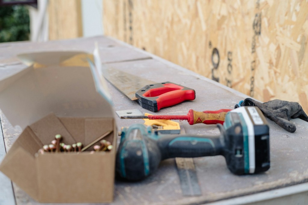 A cordless drill/driver lying on a work surface next to an open box of screws, a hand saw, a screwdriver and work gloves