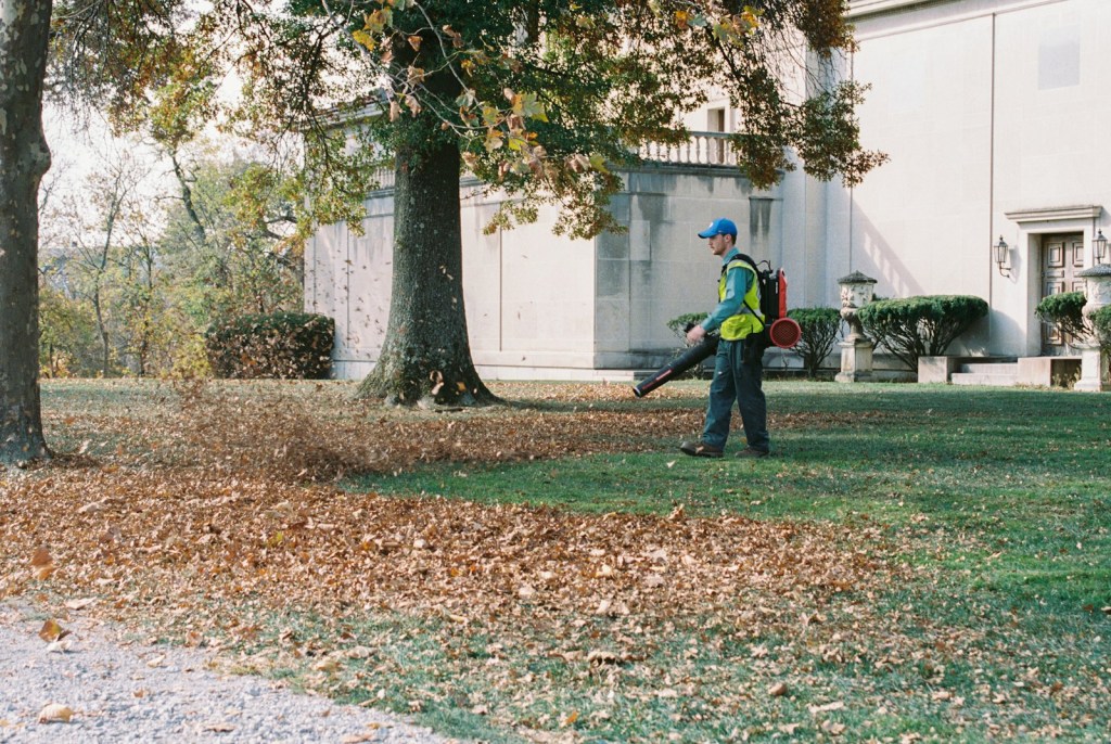 A groundskeeper in a cap and safety vest uses a backpack leaf blower to clear fallen autumn leaves from a lawn in front of a stone building
