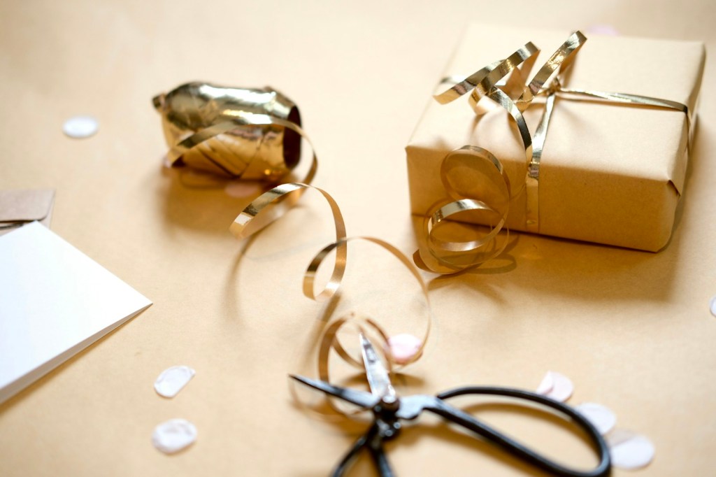A kraft-paper wrapped gift tied with gold ribbon, a spool of curling ribbon and a pair of black scissors on a tan background