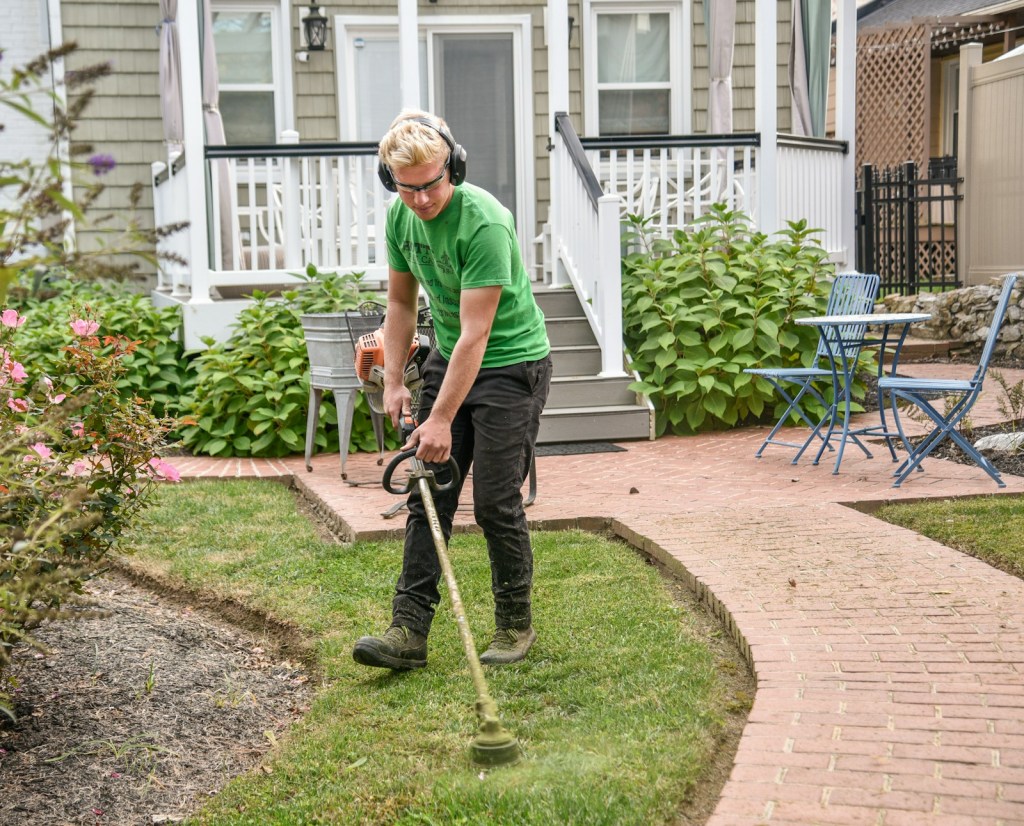 A person wearing ear protection trims the grass along a brick path with a gas-powered string trimmer