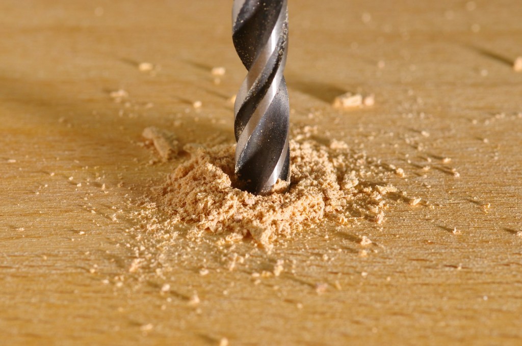 A twist drill bit boring a hole in a wooden board, with a pile of sawdust around it