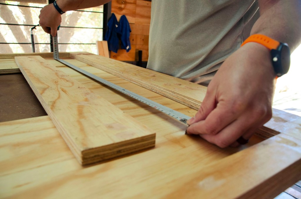 Hands holding a tape measure across a plywood board in a workshop