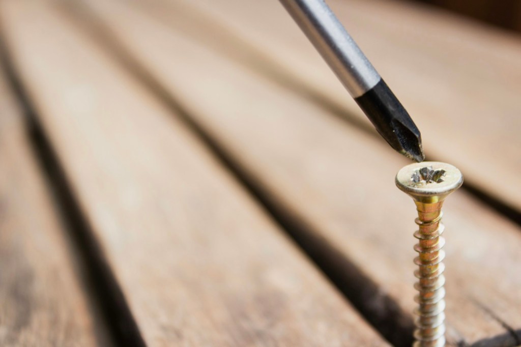 A screwdriver tip resting on the head of a gold-coloured screw standing in wooden deck boards