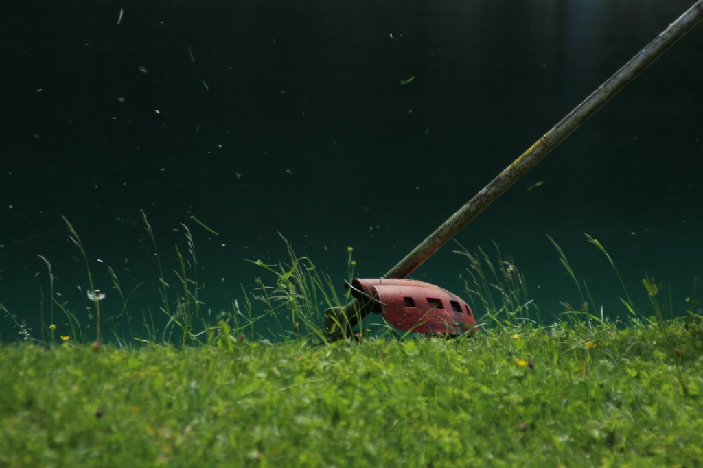 Close-up of a gas string trimmer's red guard and cutting head slicing through grass, with clippings flying against a dark green background